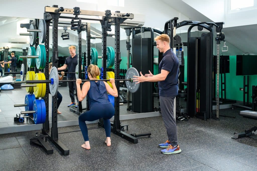 Photo of a physiotherapist coaching a woman performing a barbell squat in a well-equipped gym