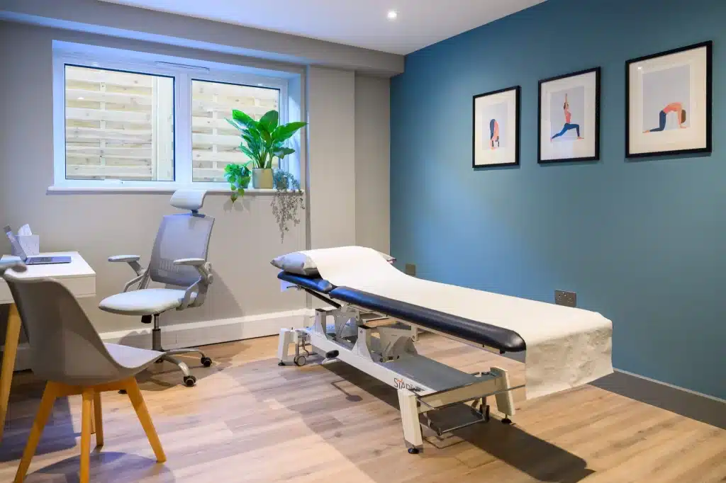 Photo of a calm physiotherapy treatment room with a treatment table, chairs, plants and framed artwork