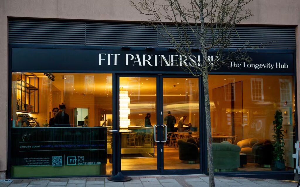 Photo of the Fit Partnership and The Longevity Hub storefront with large glass windows showing a warmly lit interior