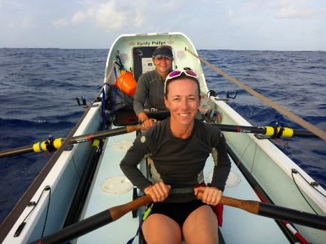 Sarah smiling at the oars of her ocean rowing boat in the Atlantic, with a crewmate sitting behind her